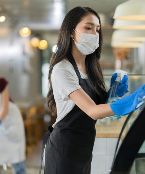 attractive positive young Asian woman staff in uniform apron spraying detergent or alcohol on counter product cabinet  wiping it with soft cloth when disinfecting cafe in ther morning safe and clean